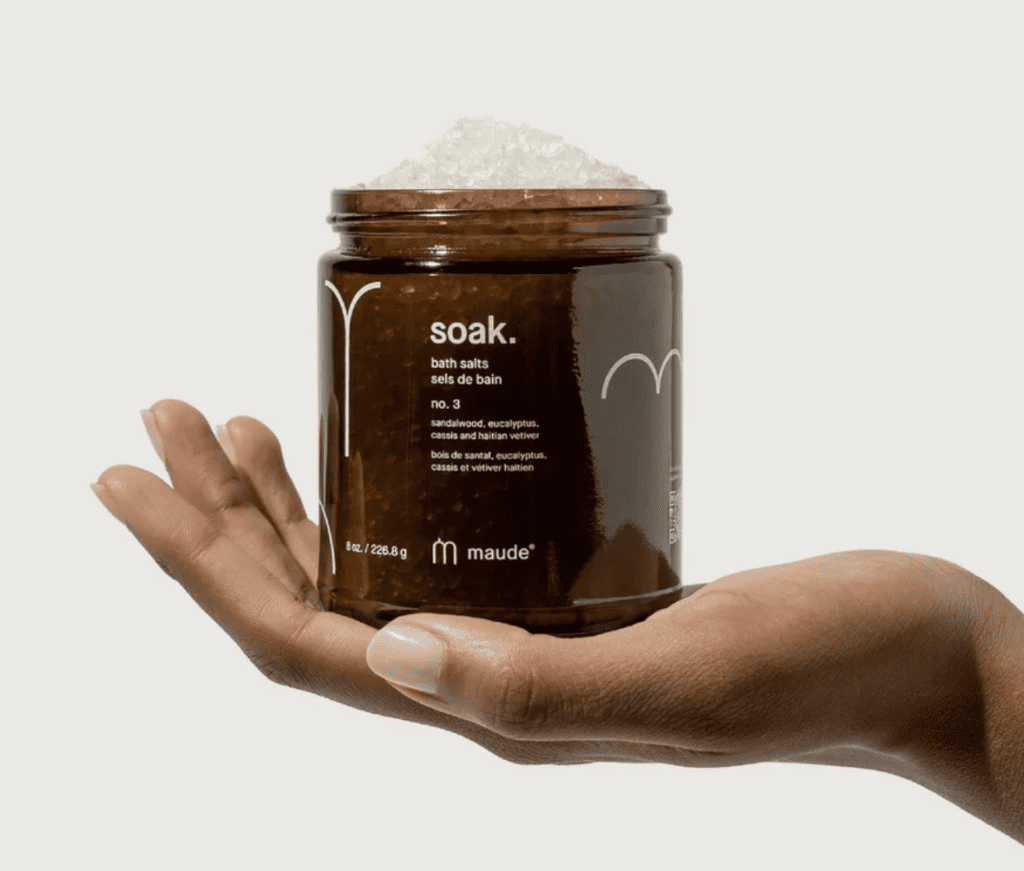 A hand holding an open container of bath salts from maude