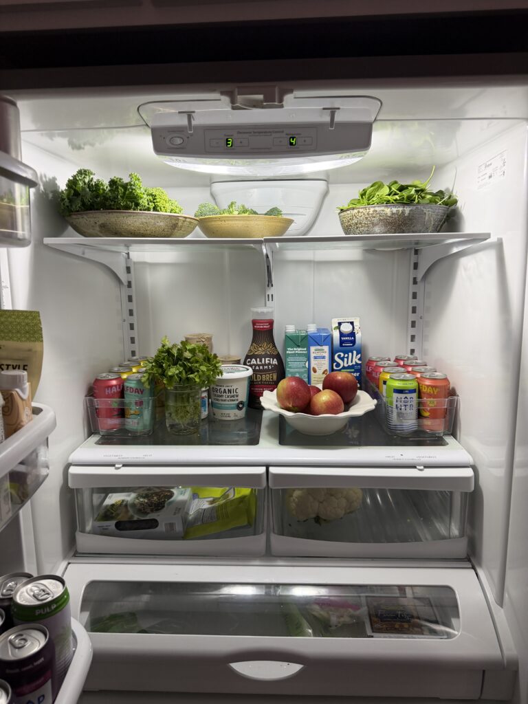 An organized fridge with fresh produce, lots of canned drinks, and a bowl of apples.