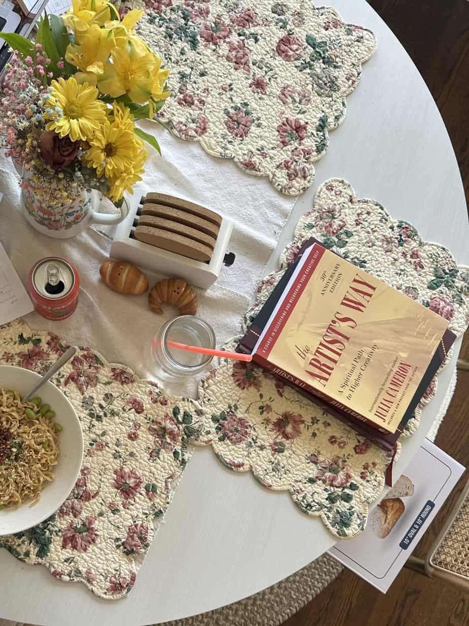 A kitchen table with water, lunch, and a journal, as well as the book 'The Artist's Way'