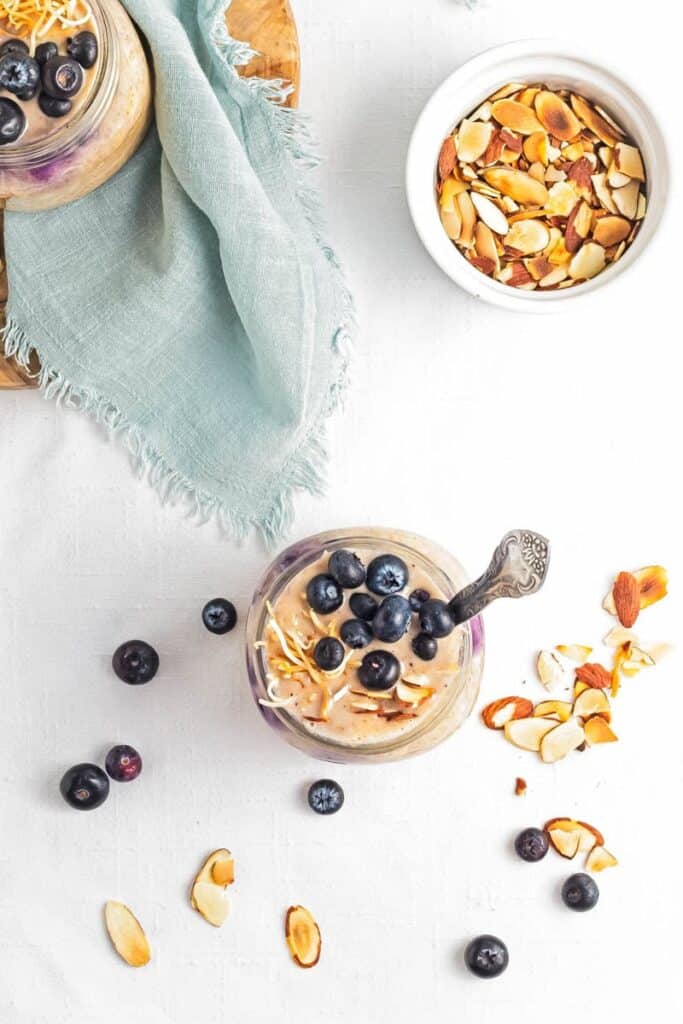 A top down shot of overnight oats topped with blueberries and toasted coconut with a bowl of almonds on the side.