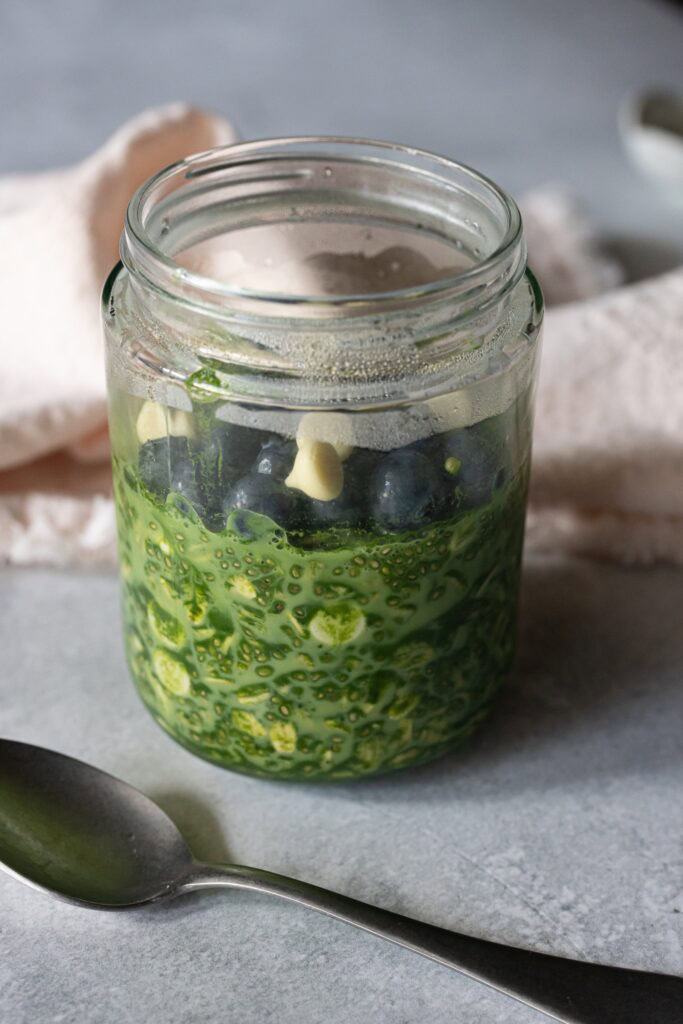 A jar of overnight oats that are green from matcha and topped with blueberries and white chocolate chips.