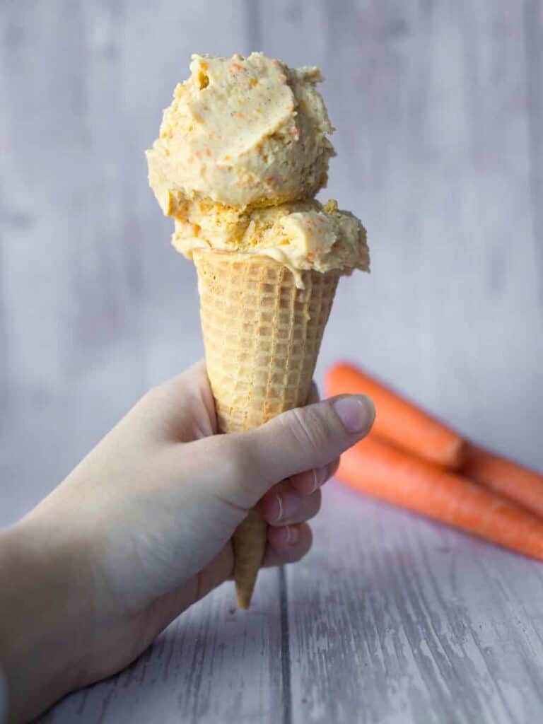 A hand holding a cone of carrot cake ice cream with carrots in the background.