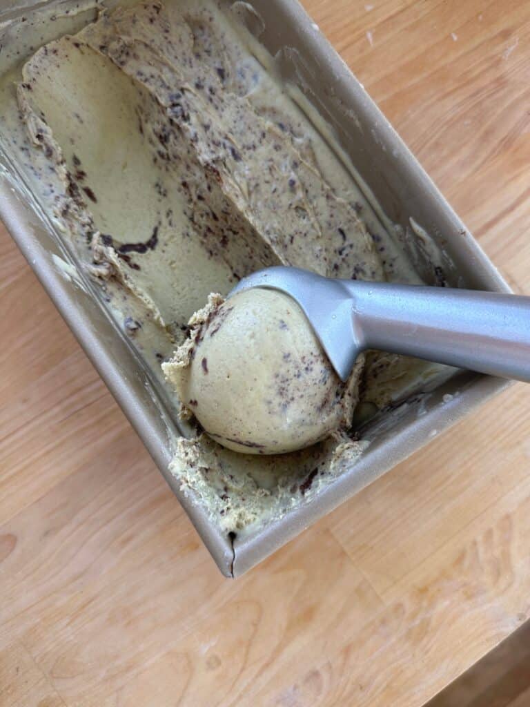 An ice cream cone with a scoop in it resting in a container of ice cream.
