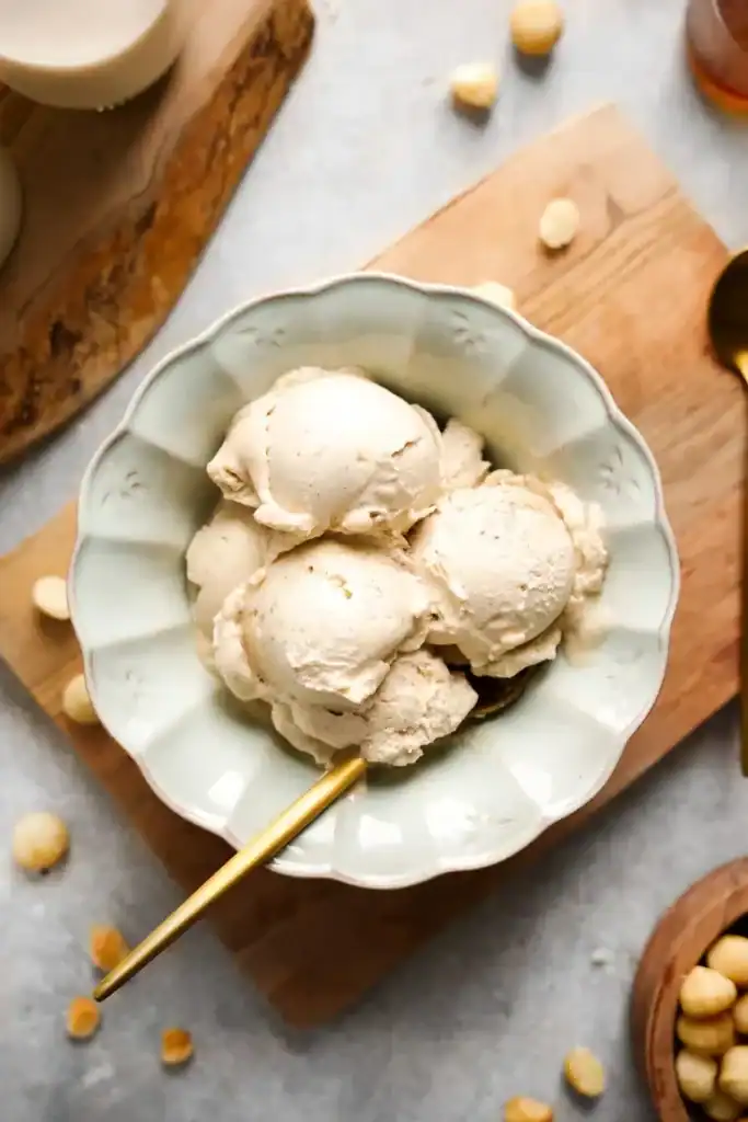 A top down shot of a bowl of macadamia nut ice cream with a gold spoon and macadamia nuts scattered around