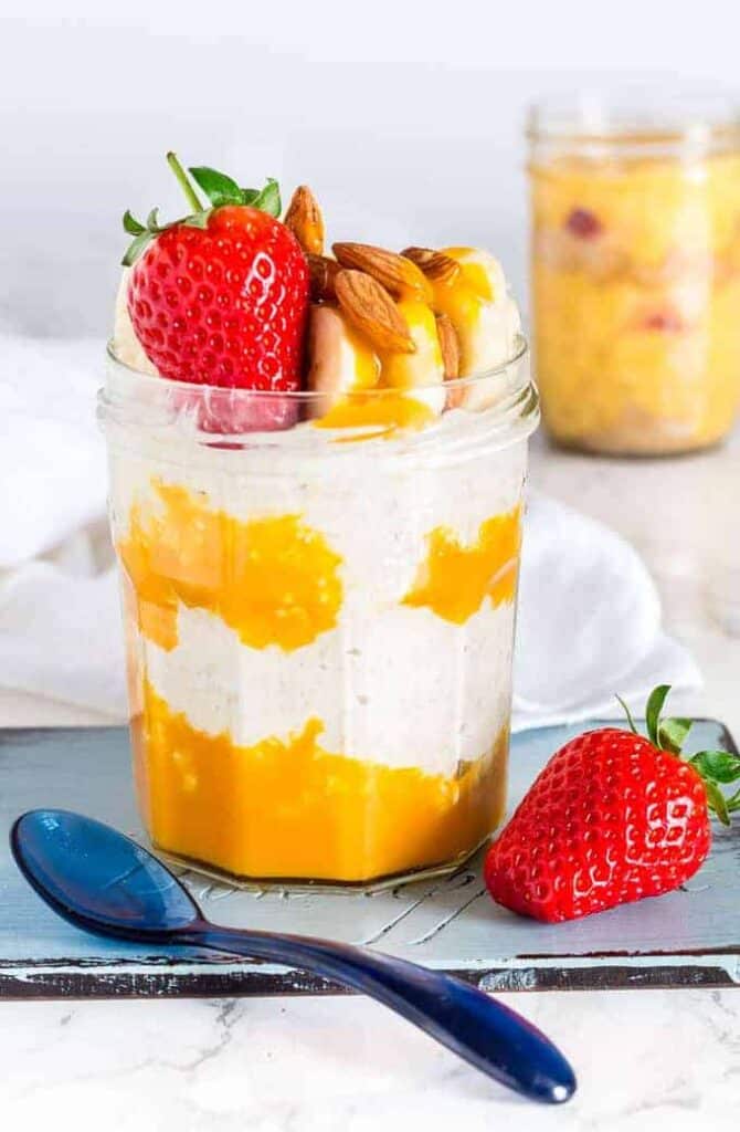 A glass with mangoes layered in between overnight oats and topped with strawberries, almonds, and bananas.