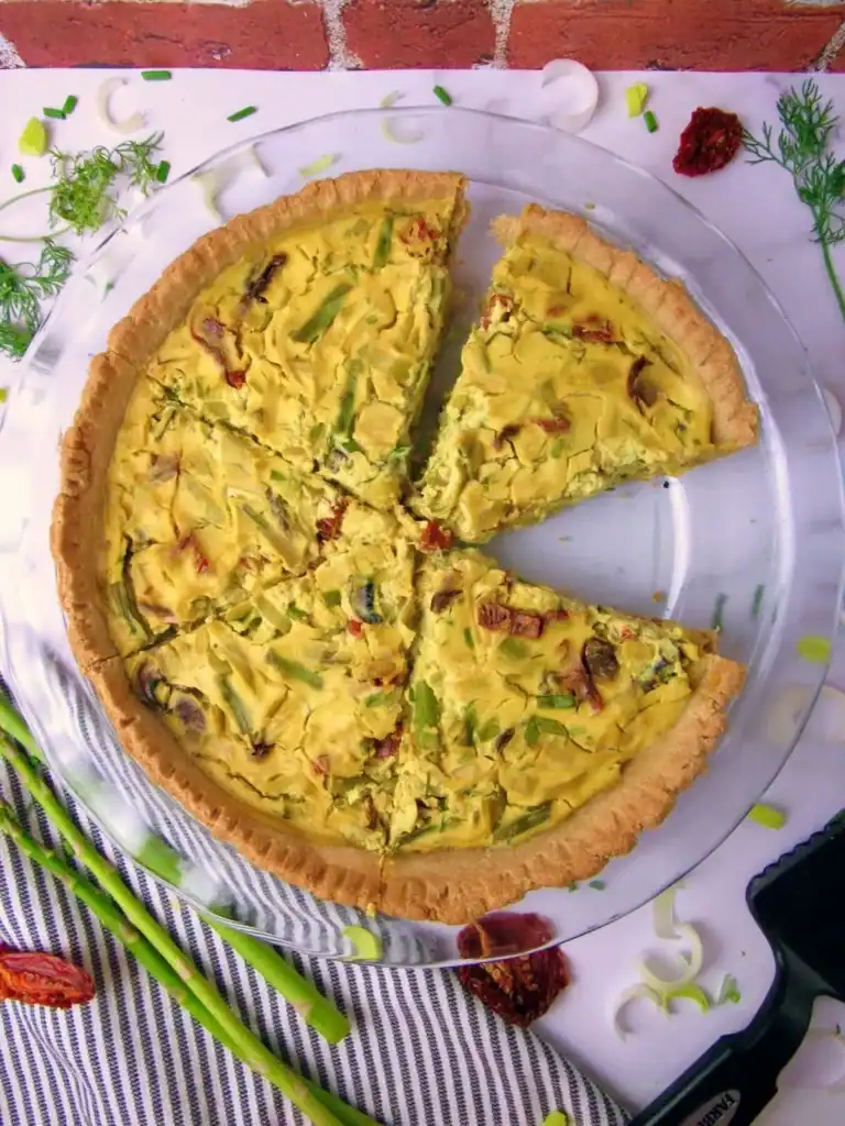 Top down shot of a vegan quiche packed with spring vegetables and surrounded by asparagus, dill, sun dried tomatoes, and scallions.