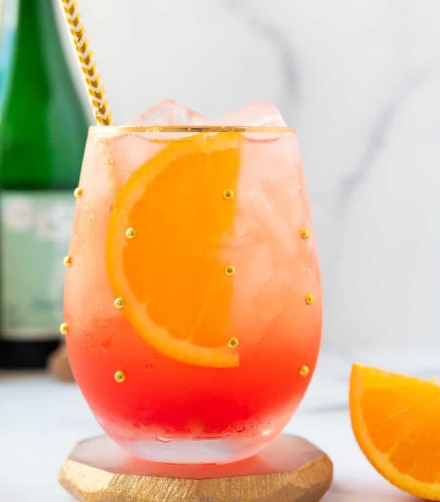 A close up of an aperol spritz mocktail in a clear glass cup with gold embellishments