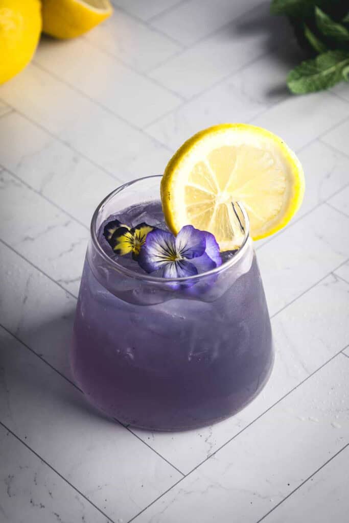 A glass filled with a purple electrolyte mocktail garnished with a lemon slice and flowers