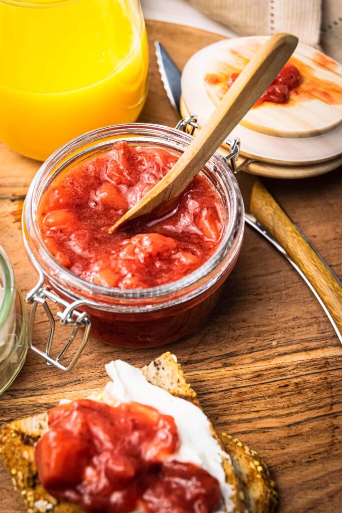 A glass jar of strawberry compote with a wooden spoon inside and surrounded by toast with compote on it