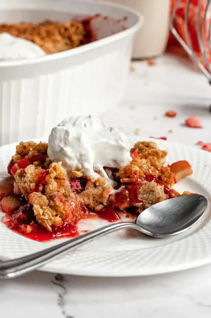 A close up of rhubarb crisp with a scoop of whipped cream on a white plate with a spoon resting nearby