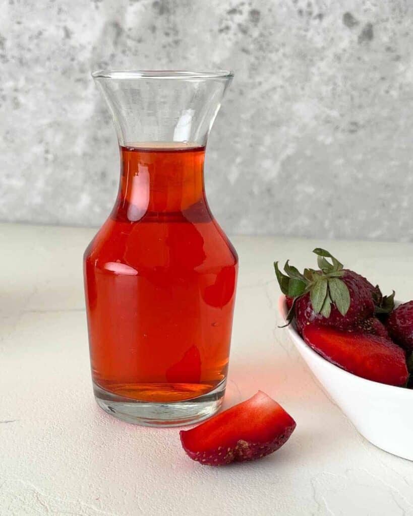 A glass filled with strawberry vinegar with a white bowl of strawberries placed nearby