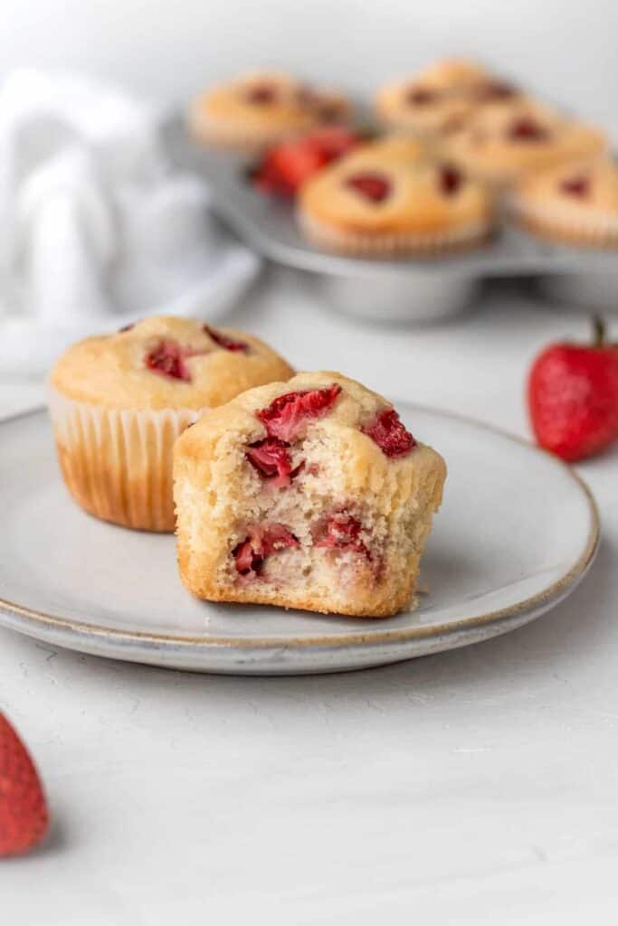 A strawberry muffin with a bite taken out of it, with a strawberry muffin in the background and a tray of strawberries in the back.