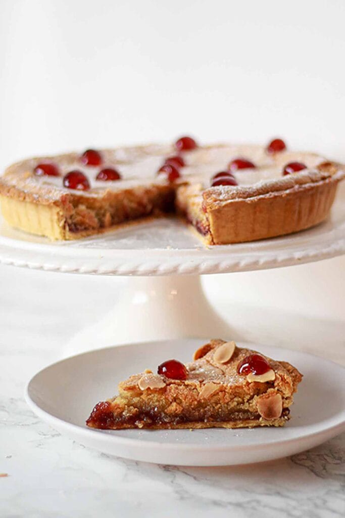 A vegan bakewell tart on a cake stand with a slice on a plate in front