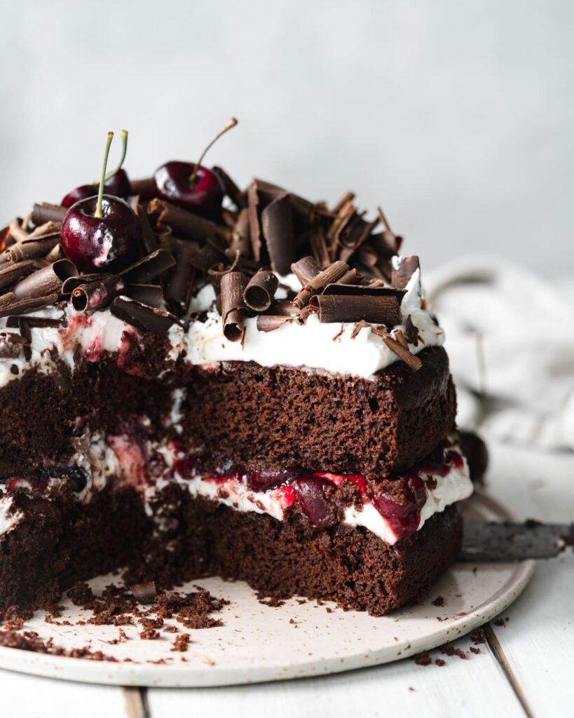 An image of a vegan black forest cake with a slice taken out and topped with chocolate and cherries